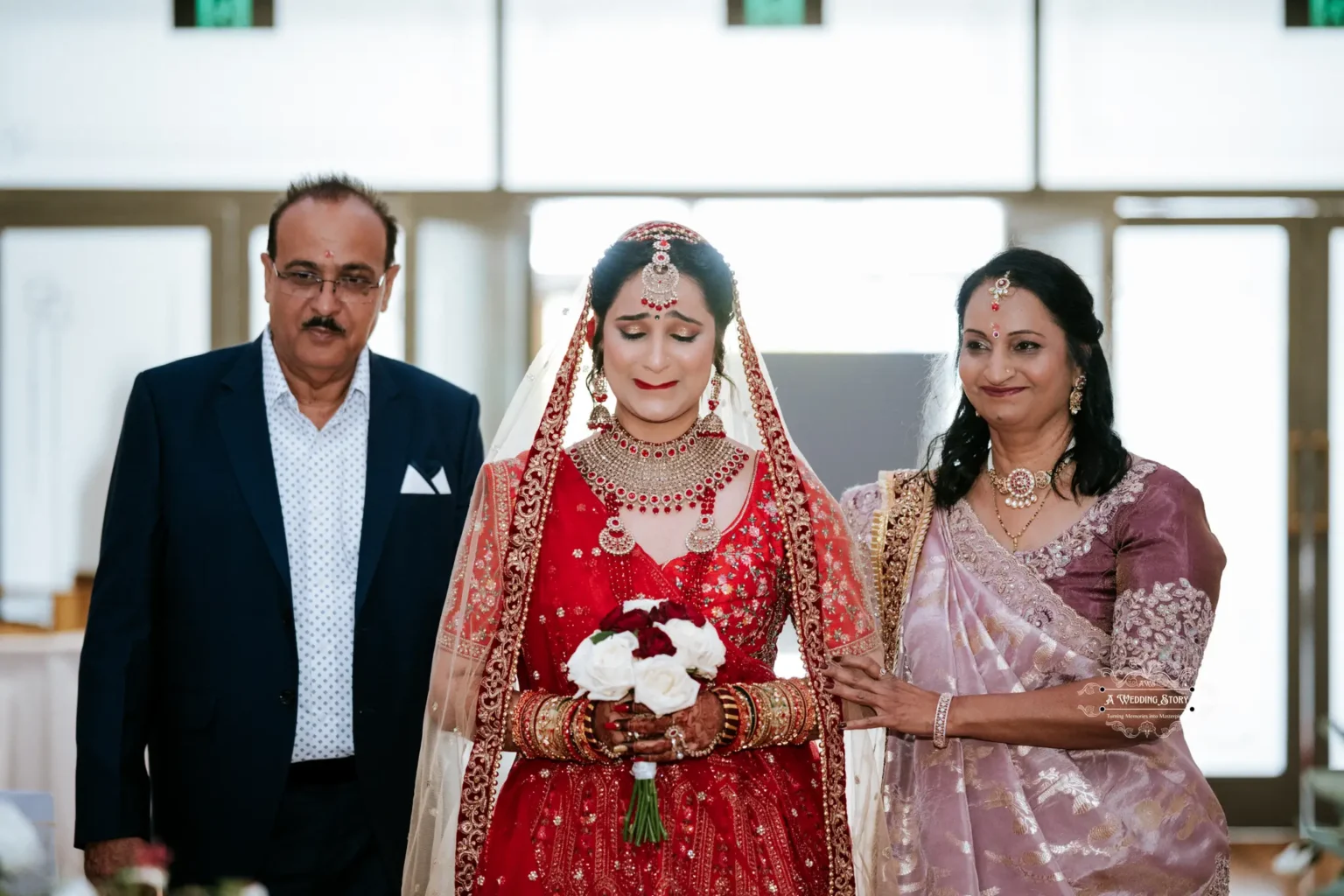 Emotional bride in traditional red attire, holding a bouquet, accompanied by her supportive parents down the aisle.