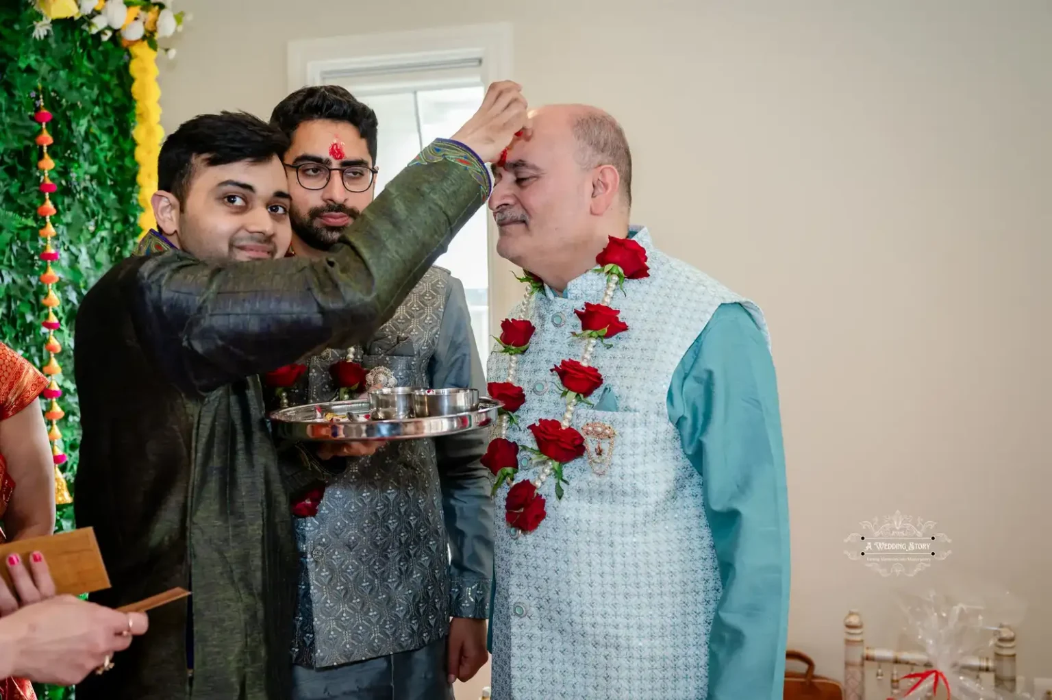 A family member applying a ceremonial tilak on the father’s forehead, while another relative holds a silver tray with sacred items, during a wedding ritual.