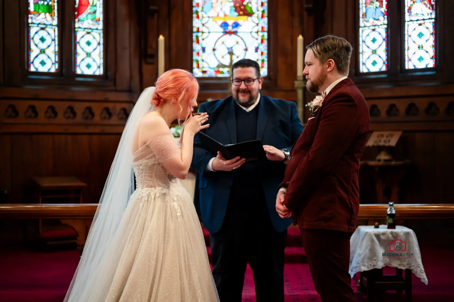 Bride and groom share their first kiss as a married couple at the altar, surrounded by family and friends in a church with stained glass windows.