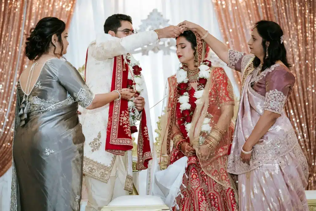 Groom applying sindoor on the bride’s forehead as part of a sacred wedding ritual, with family members assisting.