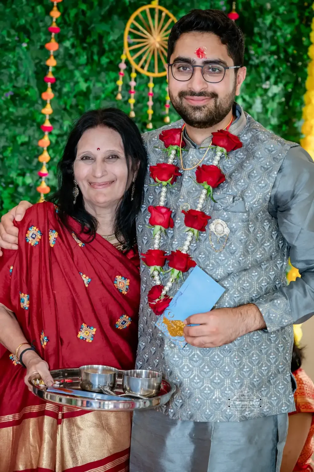 Groom in traditional attire with a garland of red roses, standing beside his mother, who holds a ceremonial tray.