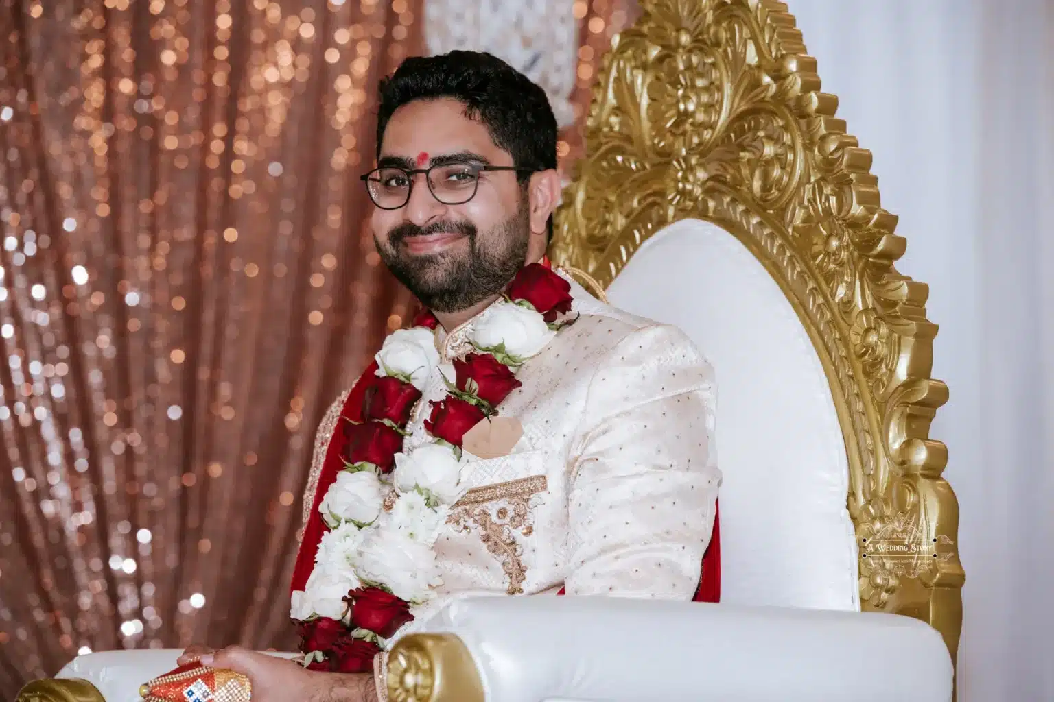 Smiling groom wearing a garland and traditional attire, seated on an ornate chair during a wedding ceremony.