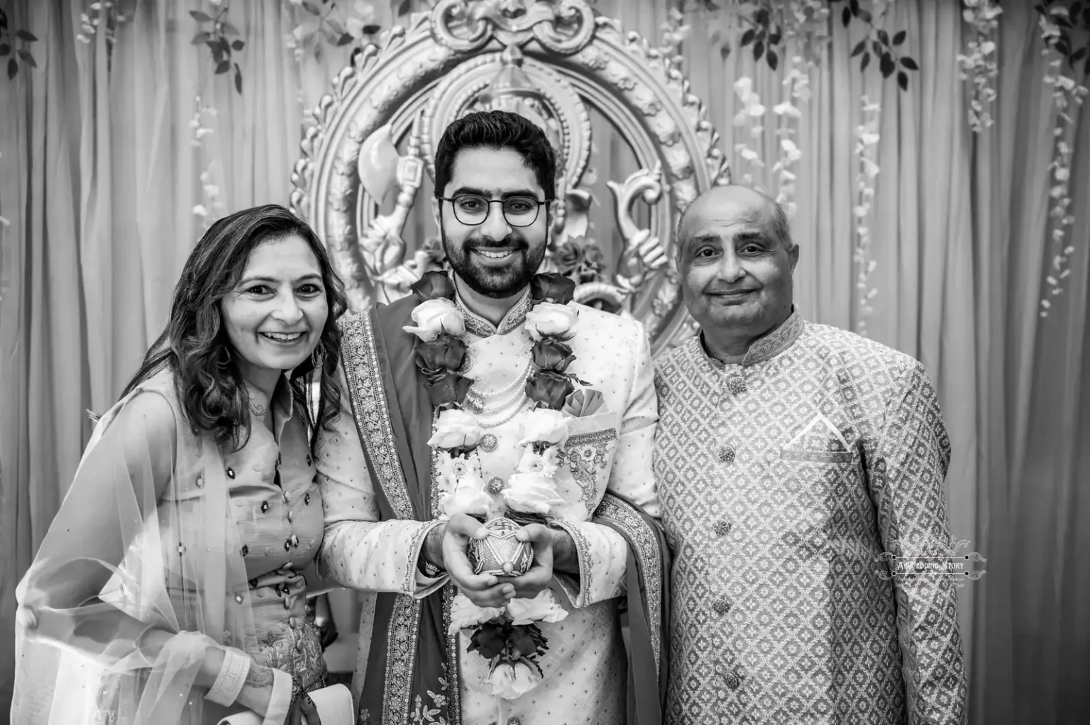 The groom stands between his parents, smiling, in a traditional Indian wedding setup with ornate decorations in Wellington.