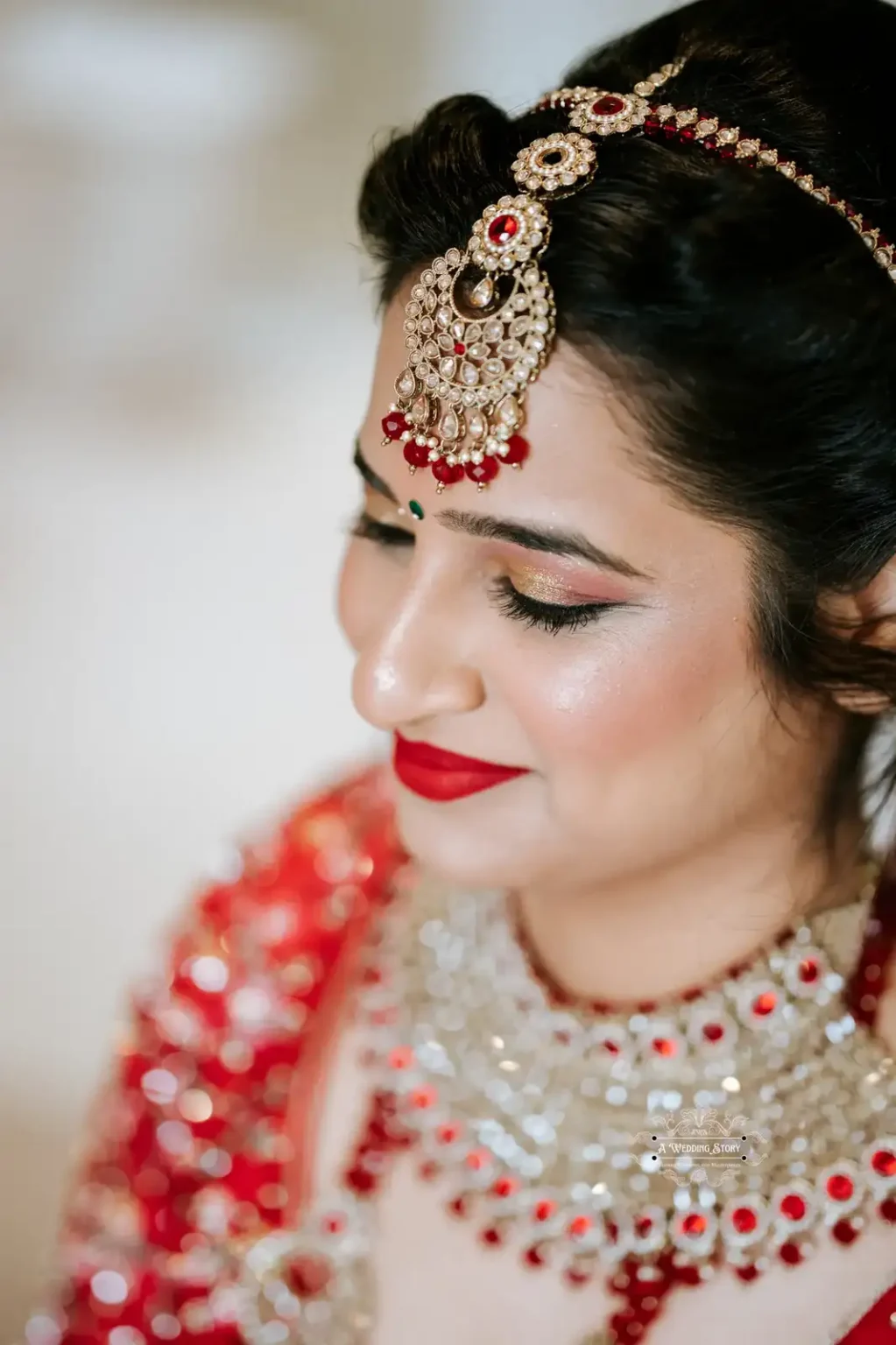 Side profile of an Indian bride in red bridal attire, wearing a gold and red maang tikka, bold red lipstick, and intricate jewelry, captured in Wellington by A Wedding Story