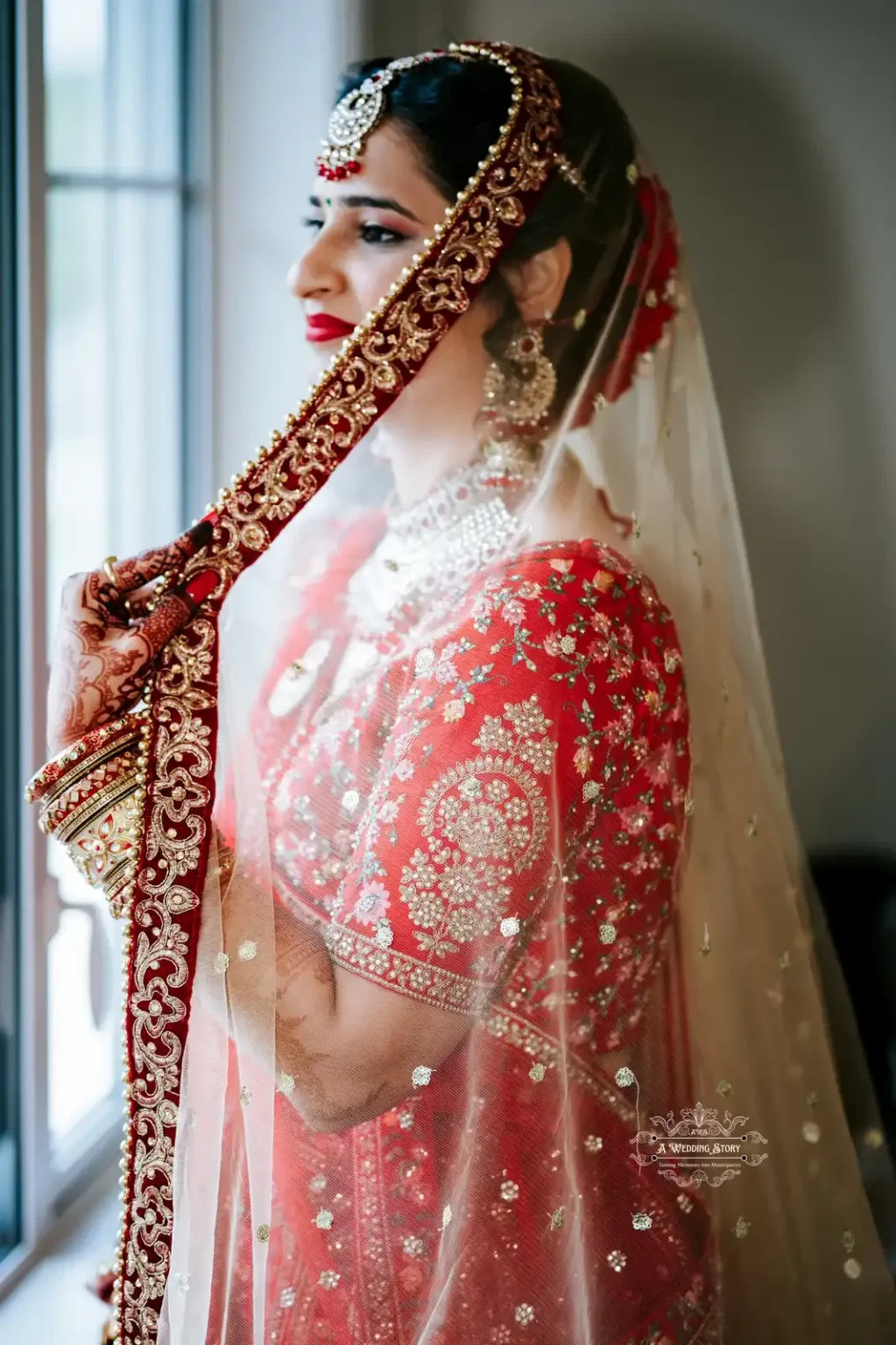 Indian bride in traditional red attire holding her veil, standing by the window, captured in Wellington by Wedding Photography.