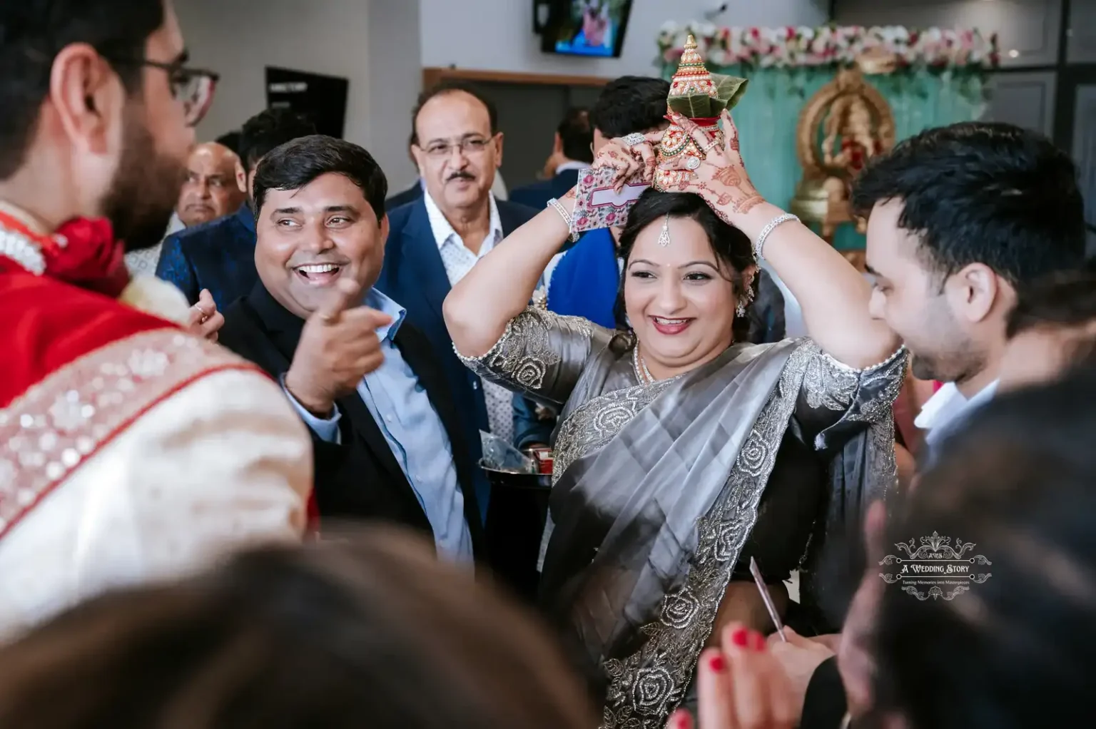 A joyful woman in a silver saree holding a traditional ornament on her head, surrounded by family and friends at a wedding ceremony in Wellington