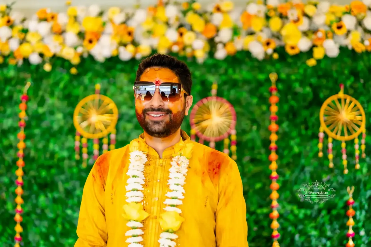 Groom wearing sunglasses and a yellow outfit with floral garlands, smiling against a vibrant floral and green backdrop during the Haldi ceremony in Wellington.