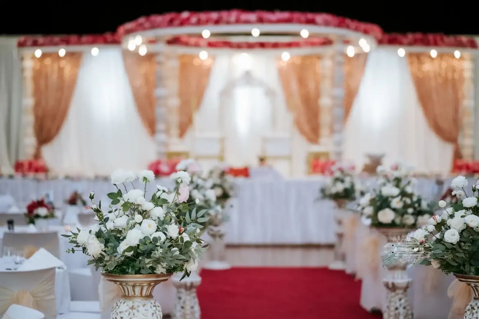 Luxurious wedding stage setup with white and gold drapes, illuminated with soft lights, and an aisle adorned with floral arrangements.