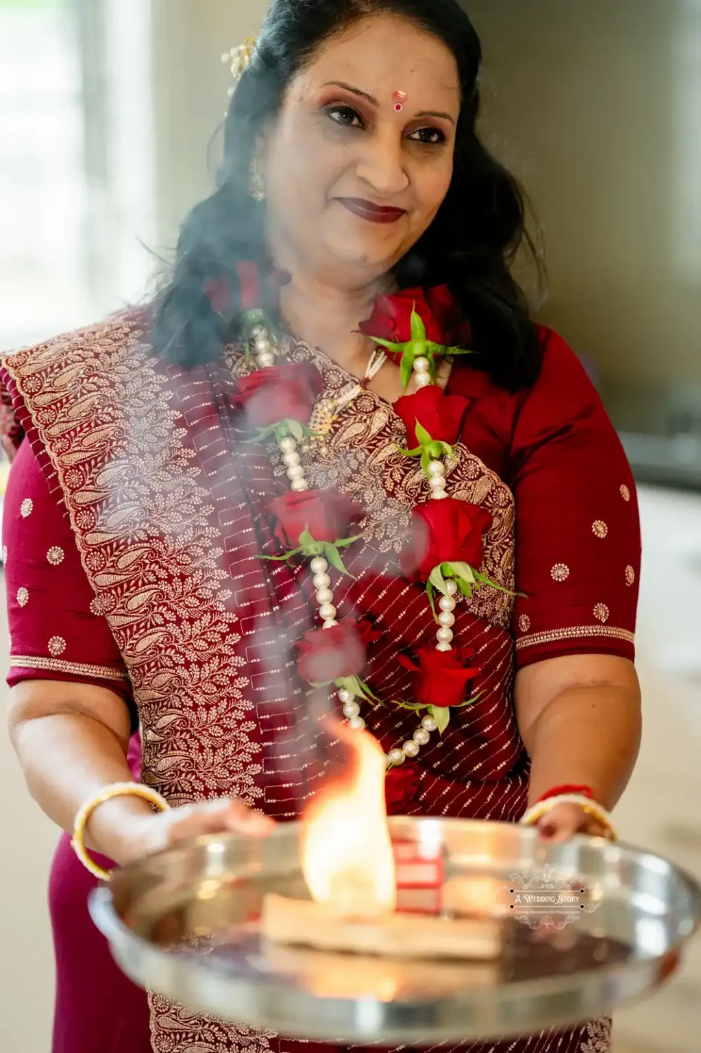 Mother dressed in a red saree, adorned with a garland of roses, performing a traditional aarti with a lit flame on a silver tray.