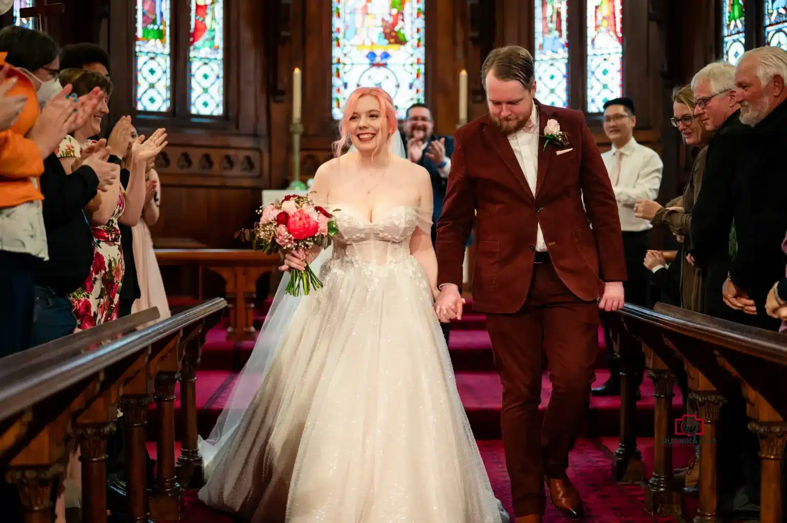 Bride and groom exiting the church after wedding ceremony, holding hands and smiling, with guests applauding on both sides of the aisle.