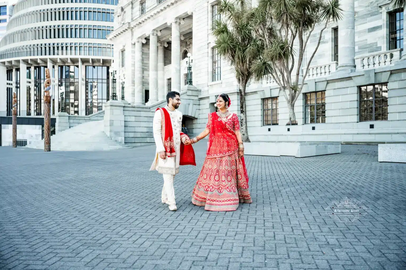 Bride and groom in traditional attire walking hand in hand outside a historic building in Wellington, New Zealand.