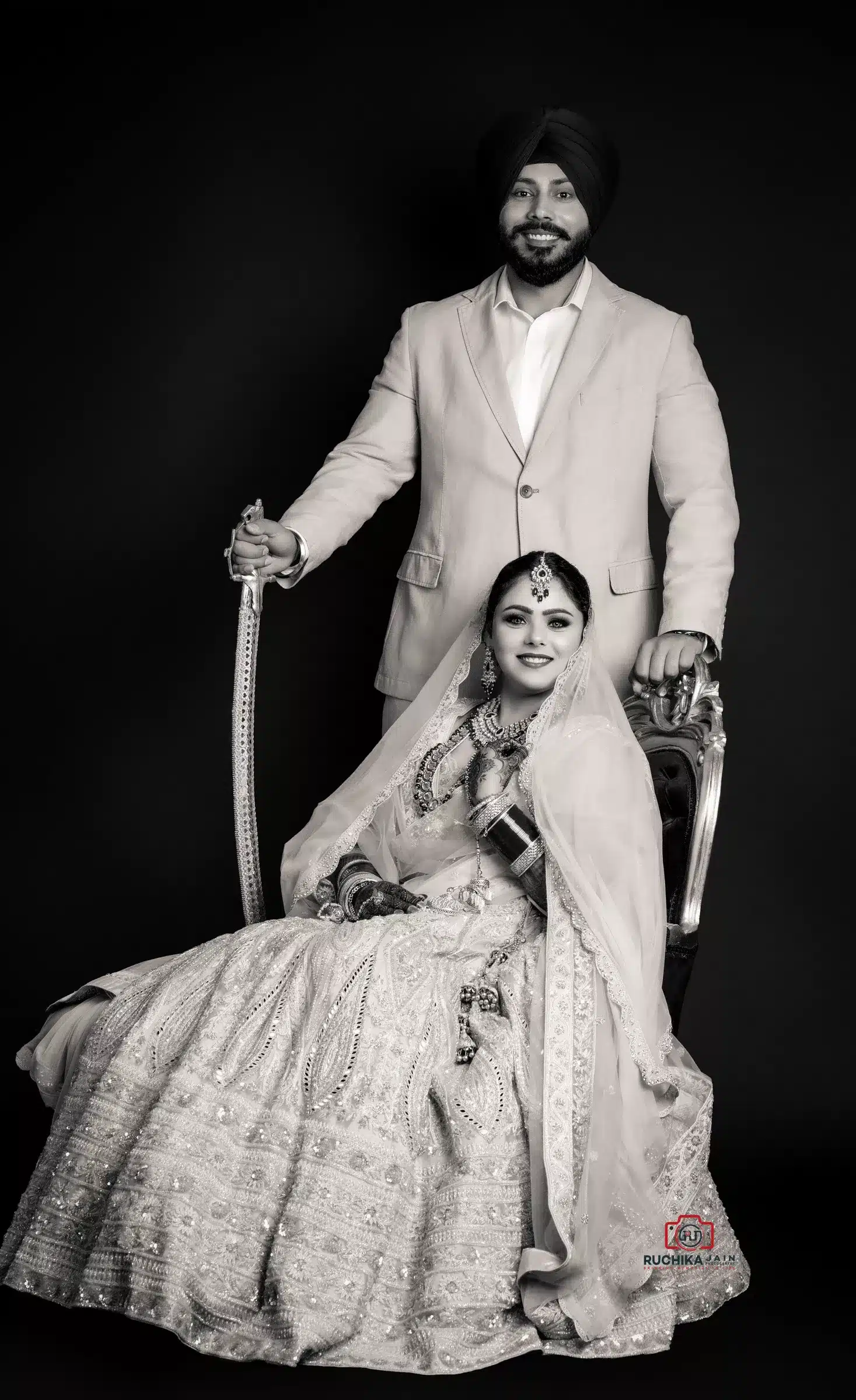 A black-and-white portrait of a Sikh wedding couple, showcasing the groom in a formal suit and the bride in a traditional lehenga, sitting on a vintage chair.