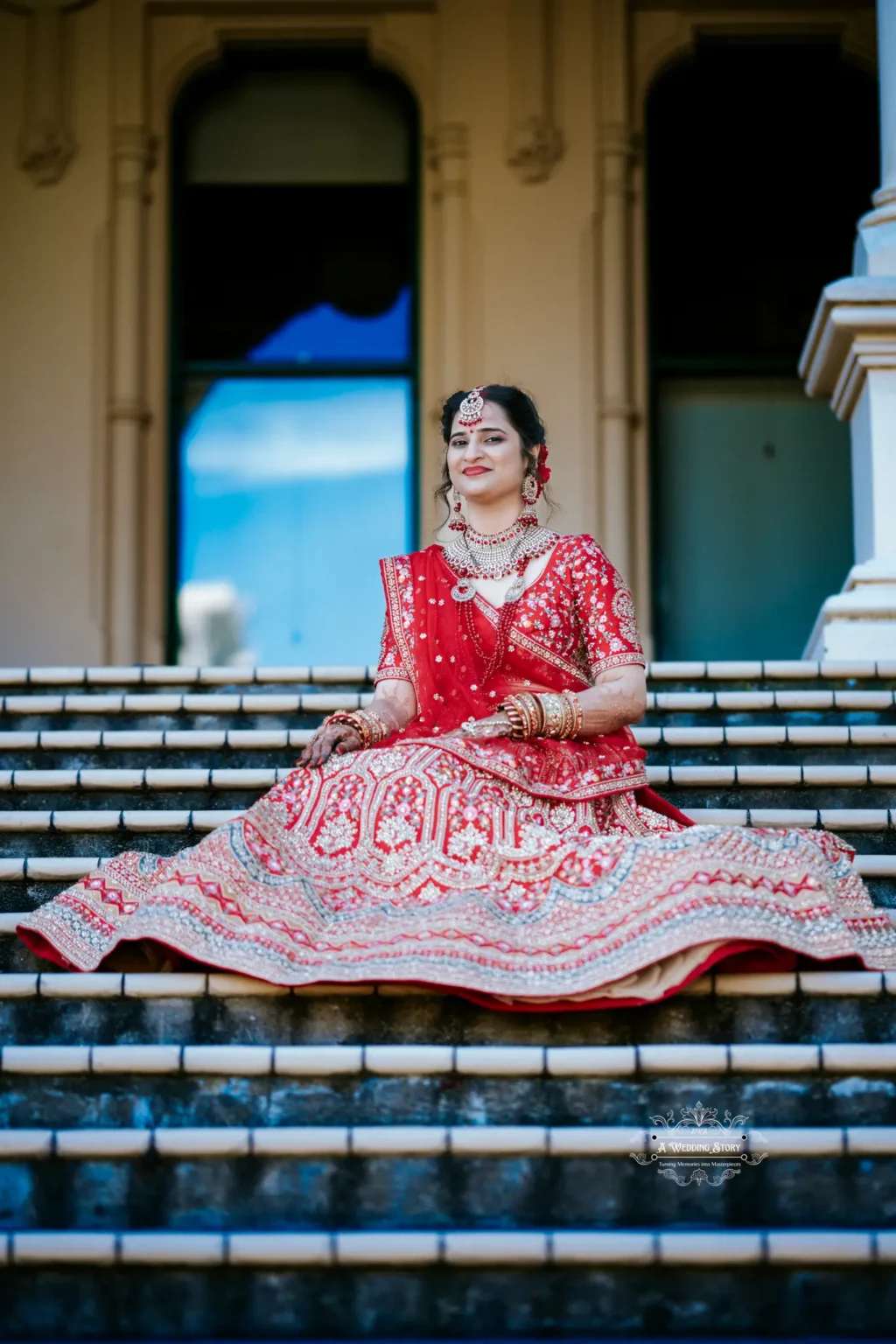 Indian bride in traditional red attire seated on steps, smiling gracefully, captured in Wellington by Wedding Photography.