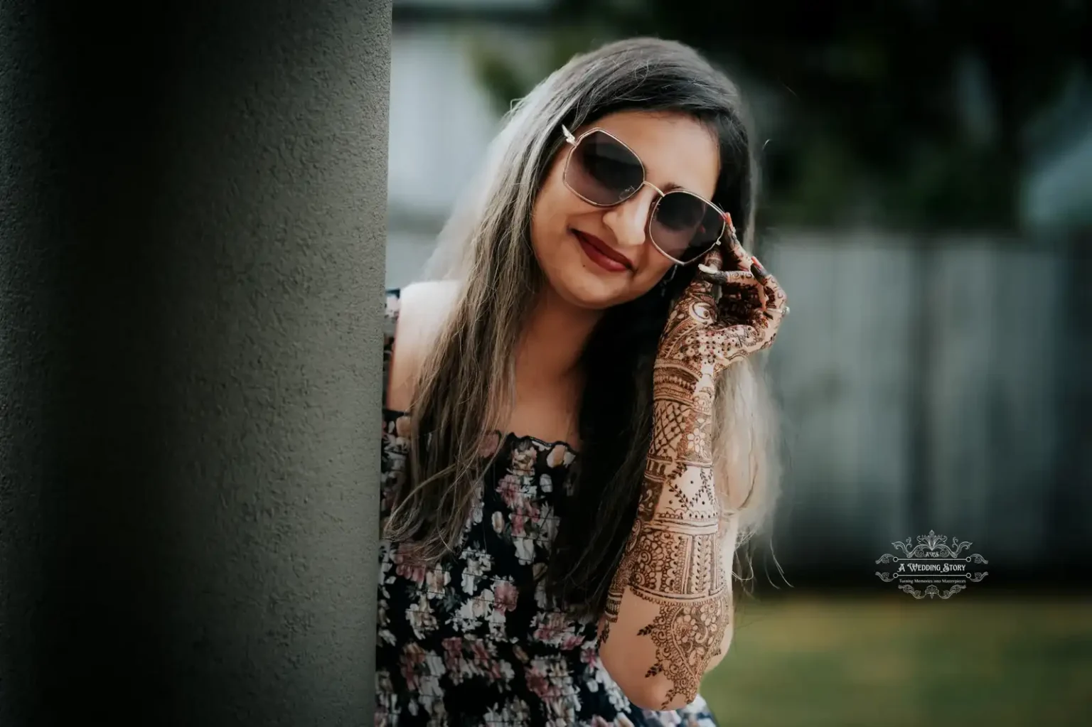 Smiling bride wearing sunglasses and showcasing intricate bridal mehndi designs on her hands, leaning playfully against a pillar during a pre-wedding celebration in Wellington by A Wedding Story