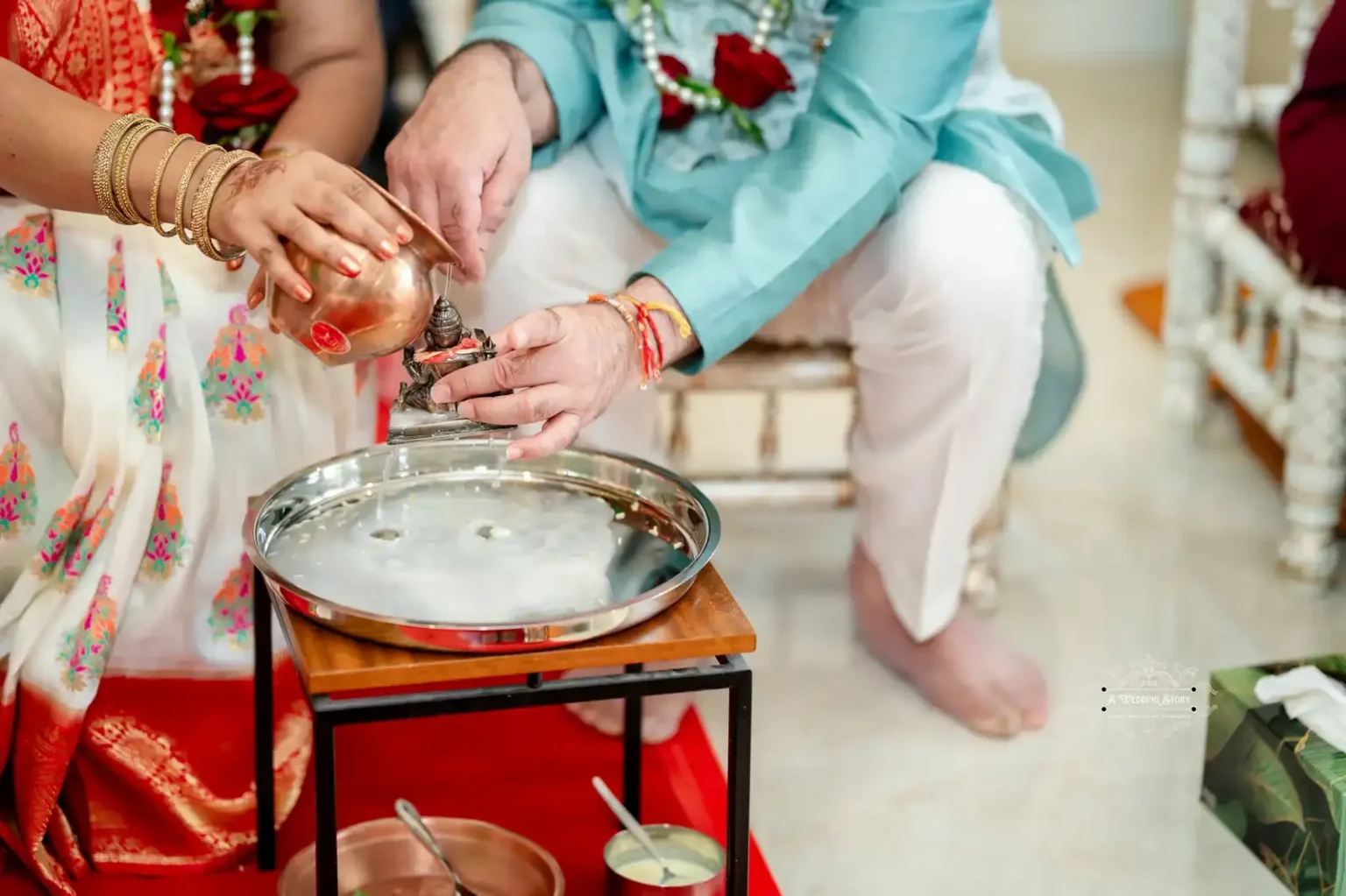 Hands performing a traditional water offering ritual during a wedding ceremony, with water poured from a copper vessel onto a deity statue over a ceremonial tray.