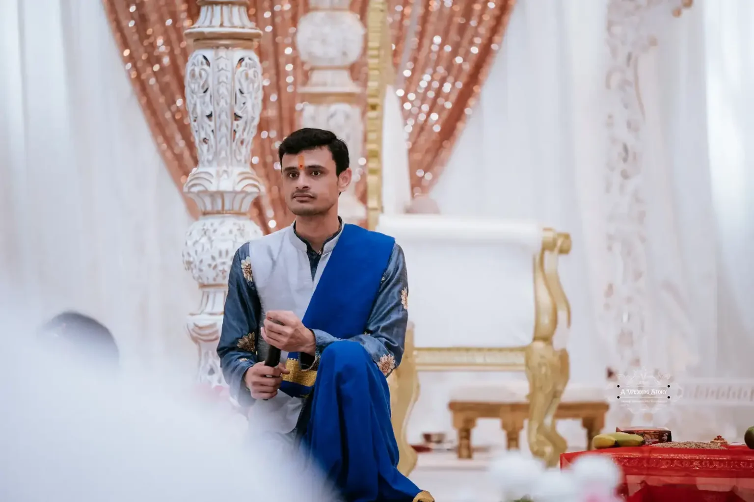 Man dressed in traditional attire holding ceremonial items, participating in an Indian wedding ritual with ornate decorations in the background.