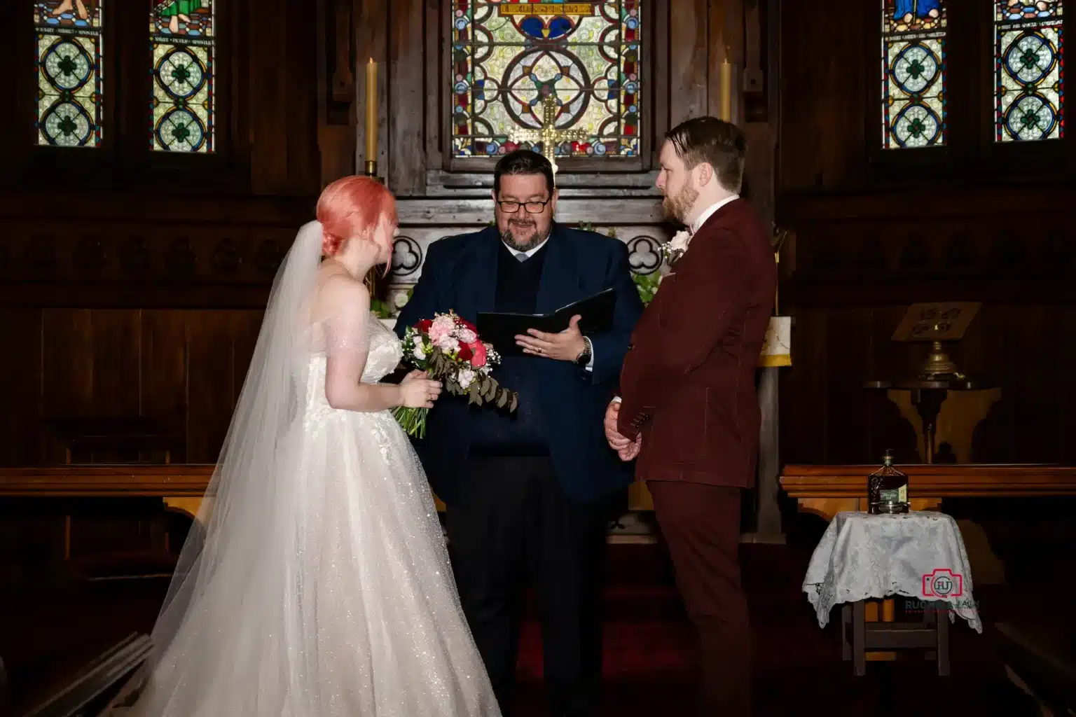 Bride and groom standing before officiant in a church, holding flowers, with stained glass windows in the background.