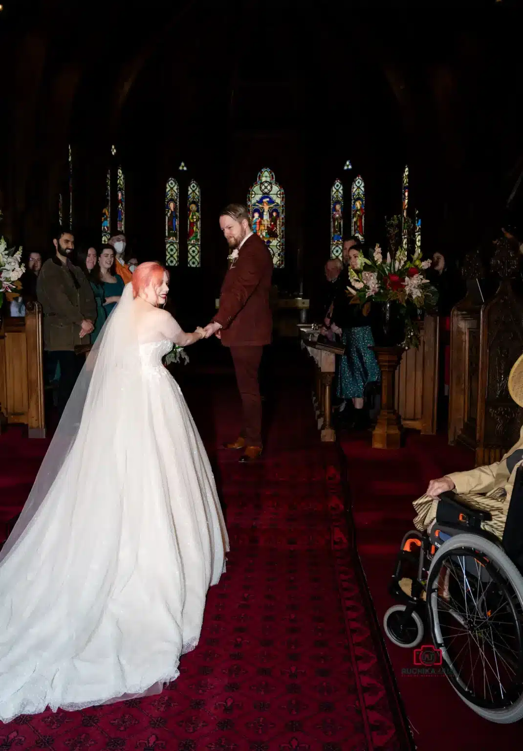 Bride and groom holding hands in a dimly lit church, surrounded by guests and stained glass windows.