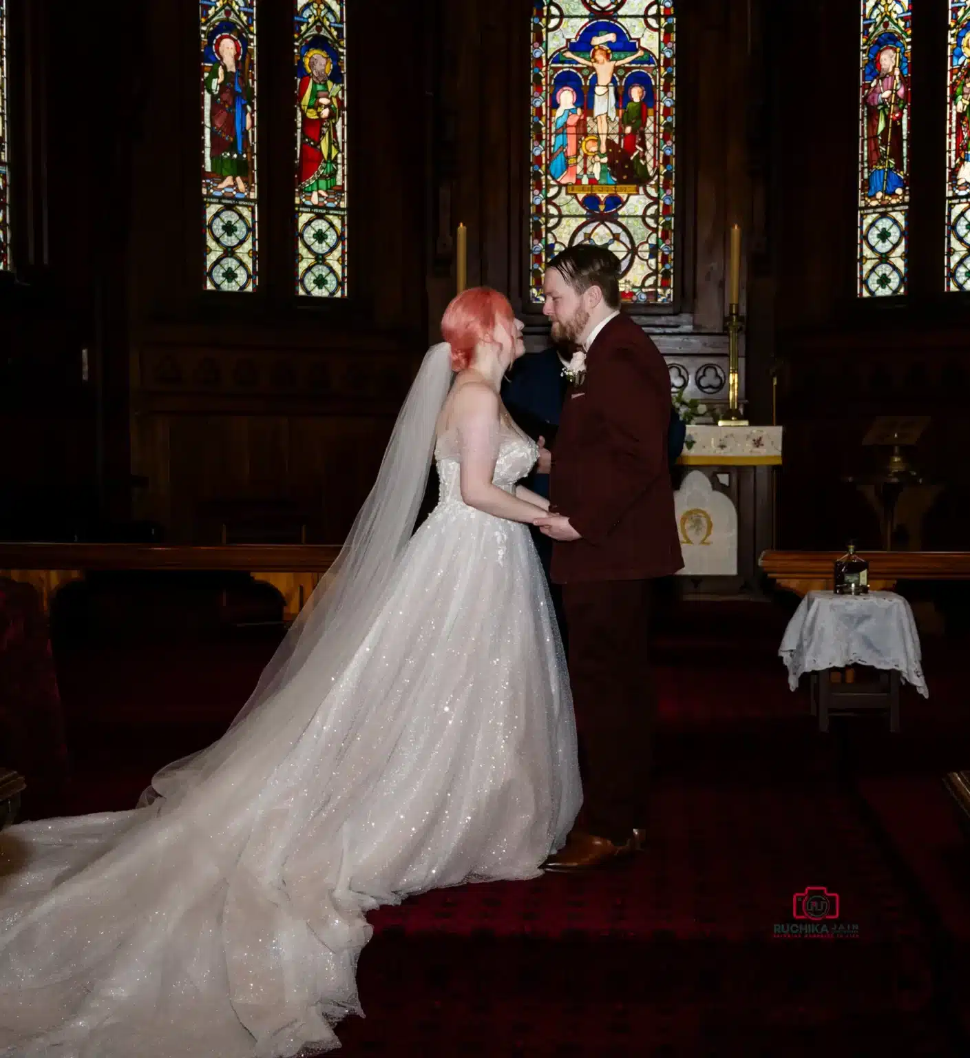 Bride and groom sharing a kiss in front of stained glass windows during a church wedding ceremony, surrounded by guests and photographers.