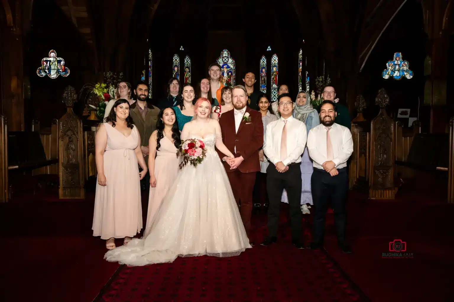 Bride, groom, and wedding party posing together in a church with stained glass windows in the background.