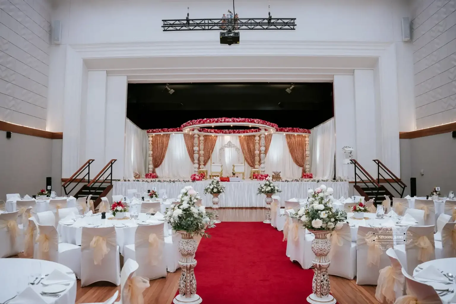 Beautifully decorated Indian wedding reception venue with a grand stage, white and gold theme, floral arrangements, and a red carpet captured in Wellington by A Wedding Story