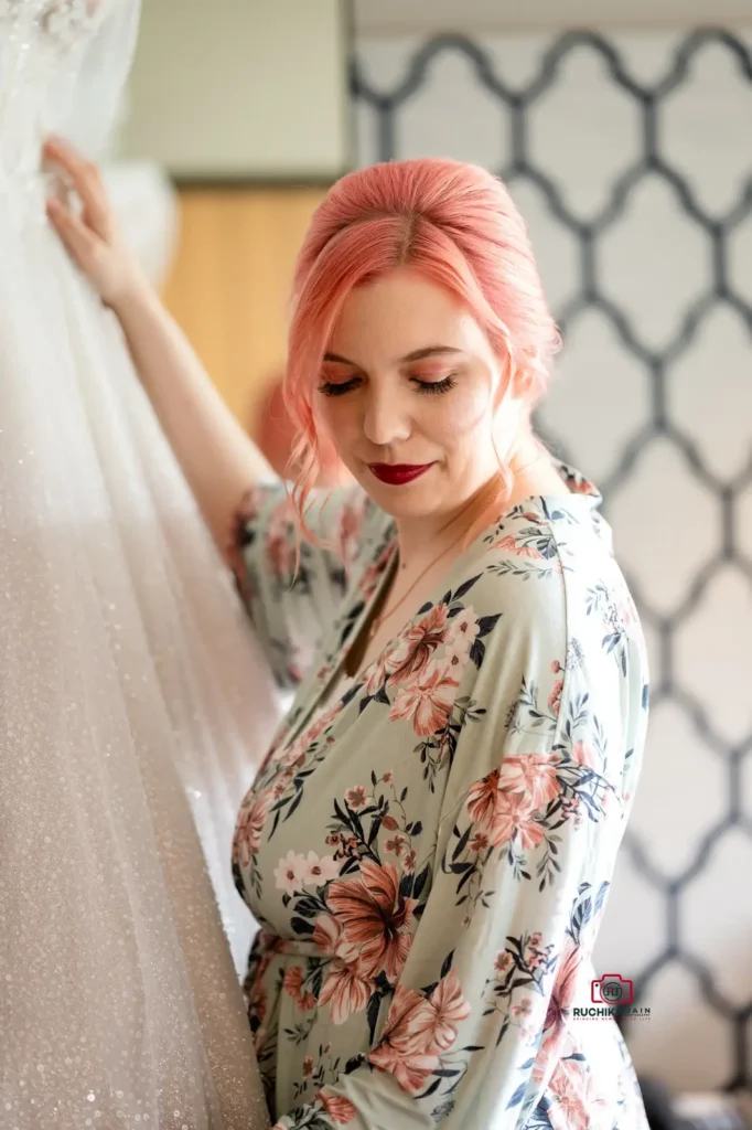 A bride with pink hair in a floral robe looking lovingly at her wedding gown before the ceremony