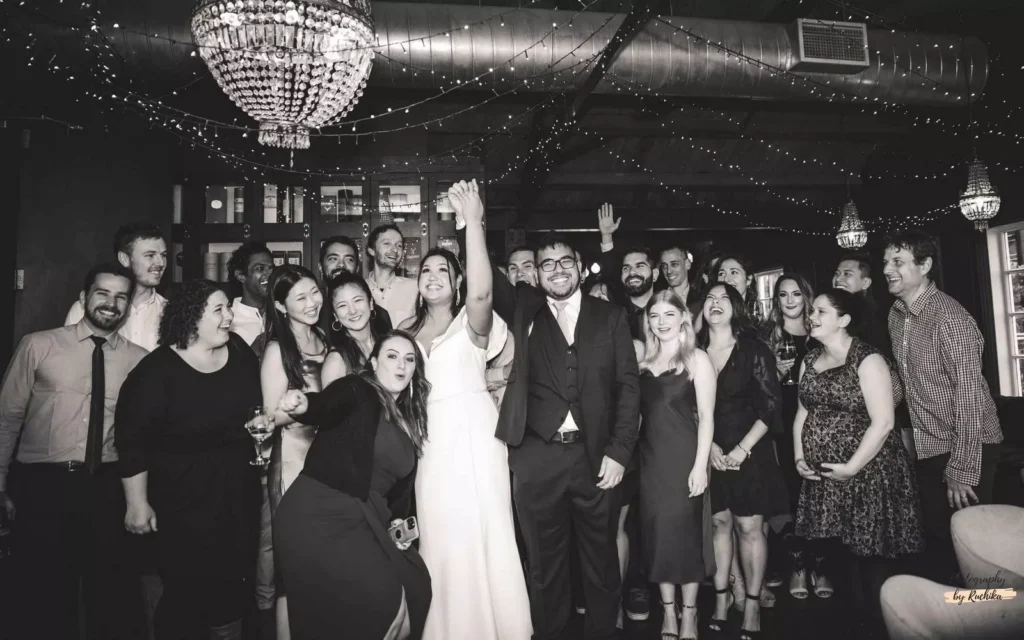 Bride and groom celebrating with friends and family at a wedding reception, all smiling and posing under string lights