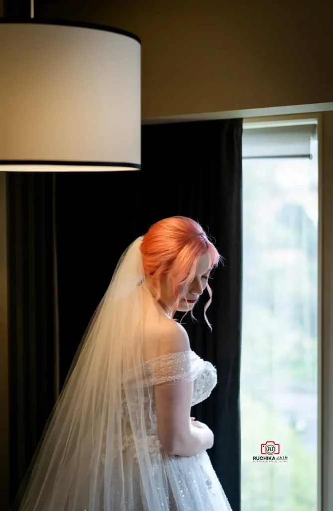 A stunning bride with pink hair gazes down thoughtfully by a window, wearing a shimmering off-the-shoulder wedding gown and a flowing veil.