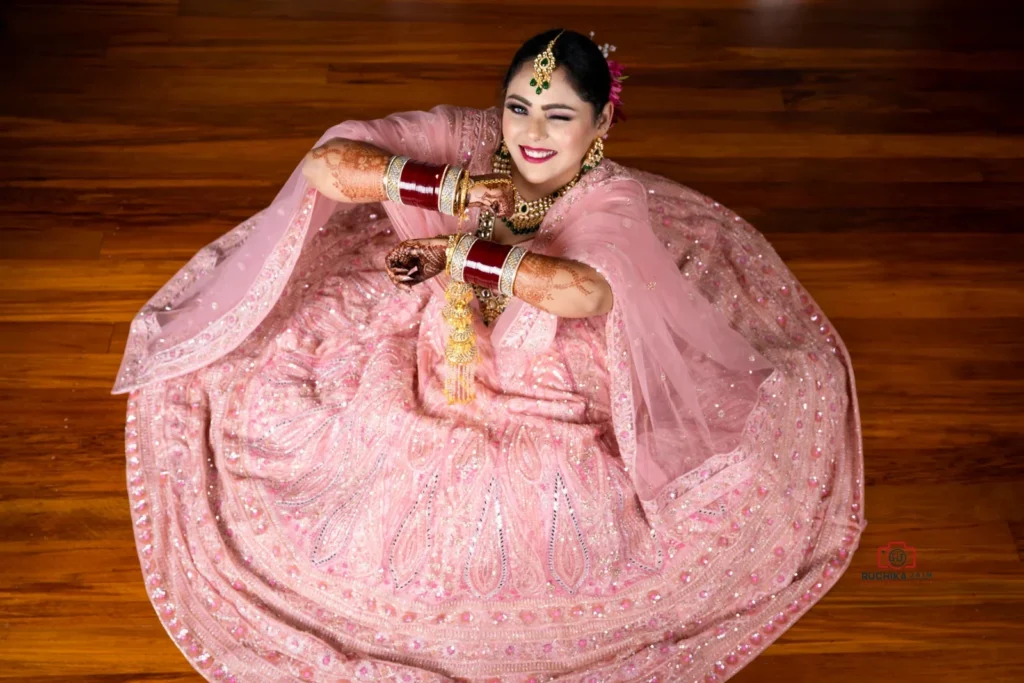 Bride in pink lehenga sitting on the floor, smiling and winking with folded arms