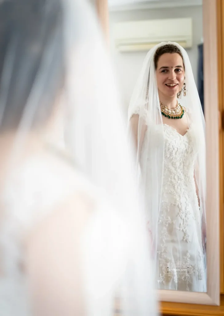 A radiant bride, wearing an elegant lace gown and a delicate veil, smiles warmly as she admires her reflection in the mirror