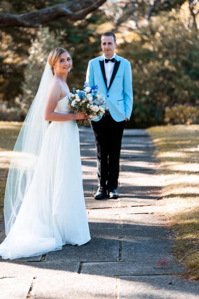 Bride Julia in a white wedding gown holding a bouquet of blue and white flowers, smiling softly while the groom, Liam, dressed in a sharp light blue tuxedo jacket with black accents, stands in the background on a serene garden pathway.