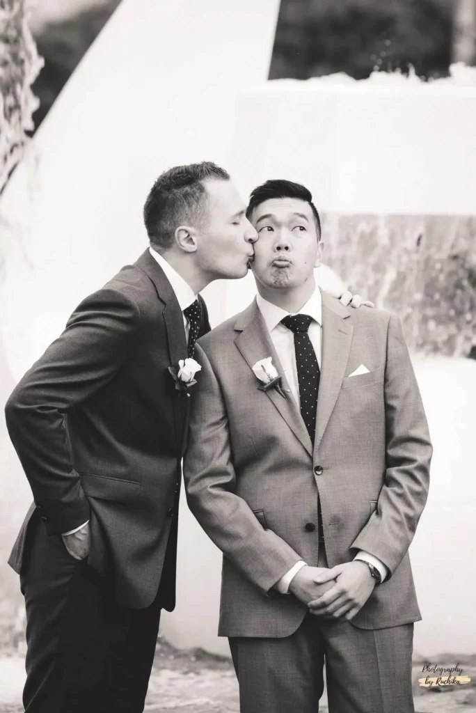 A fun and candid black-and-white photo of two grooms sharing a playful kiss during their wedding celebration in Wellington, New Zealand.