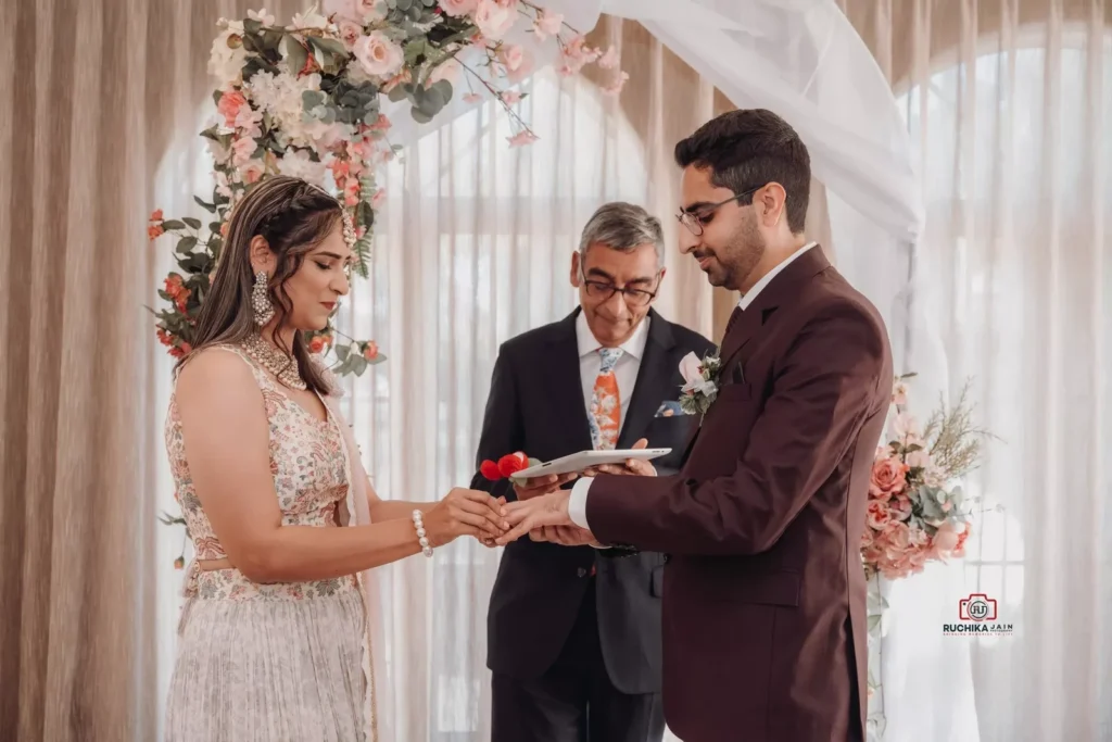 Bride and groom exchange wedding rings during an intimate ceremony, with a floral backdrop and officiant presiding