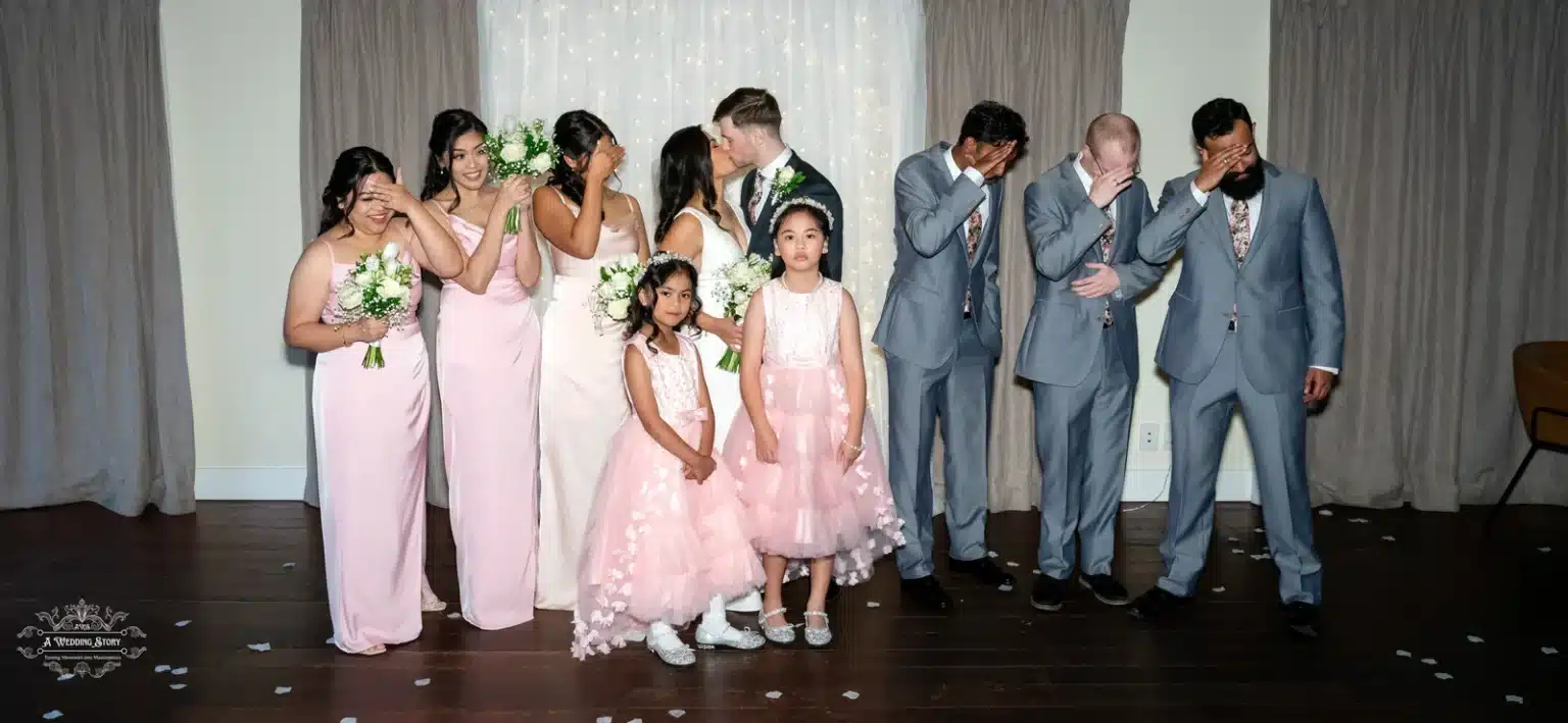 The bridal party humorously reacts to the bride and groom sharing a kiss, while two flower girls pose for the photo.