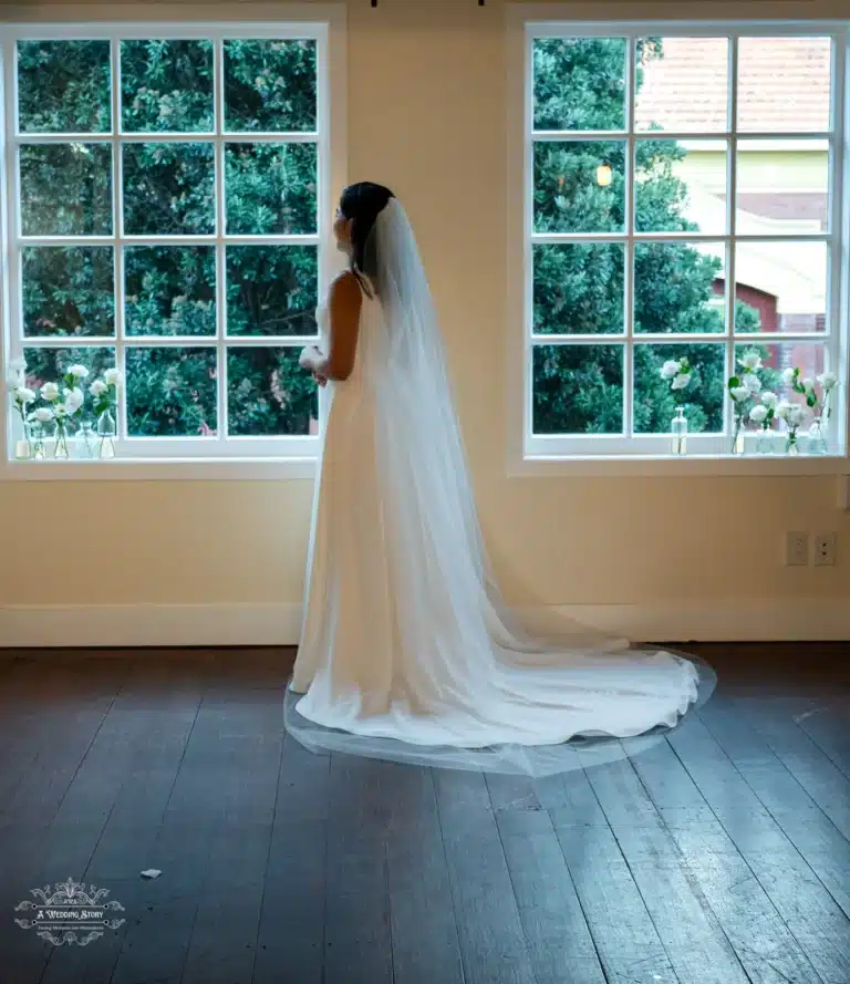 A bride in a white gown with a flowing train, standing gracefully by a window at a Wellington wedding venue.