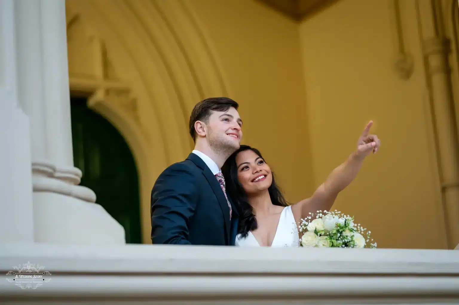 Bride and groom standing on a historic balcony, gazing and pointing towards the horizon with joyful smiles.