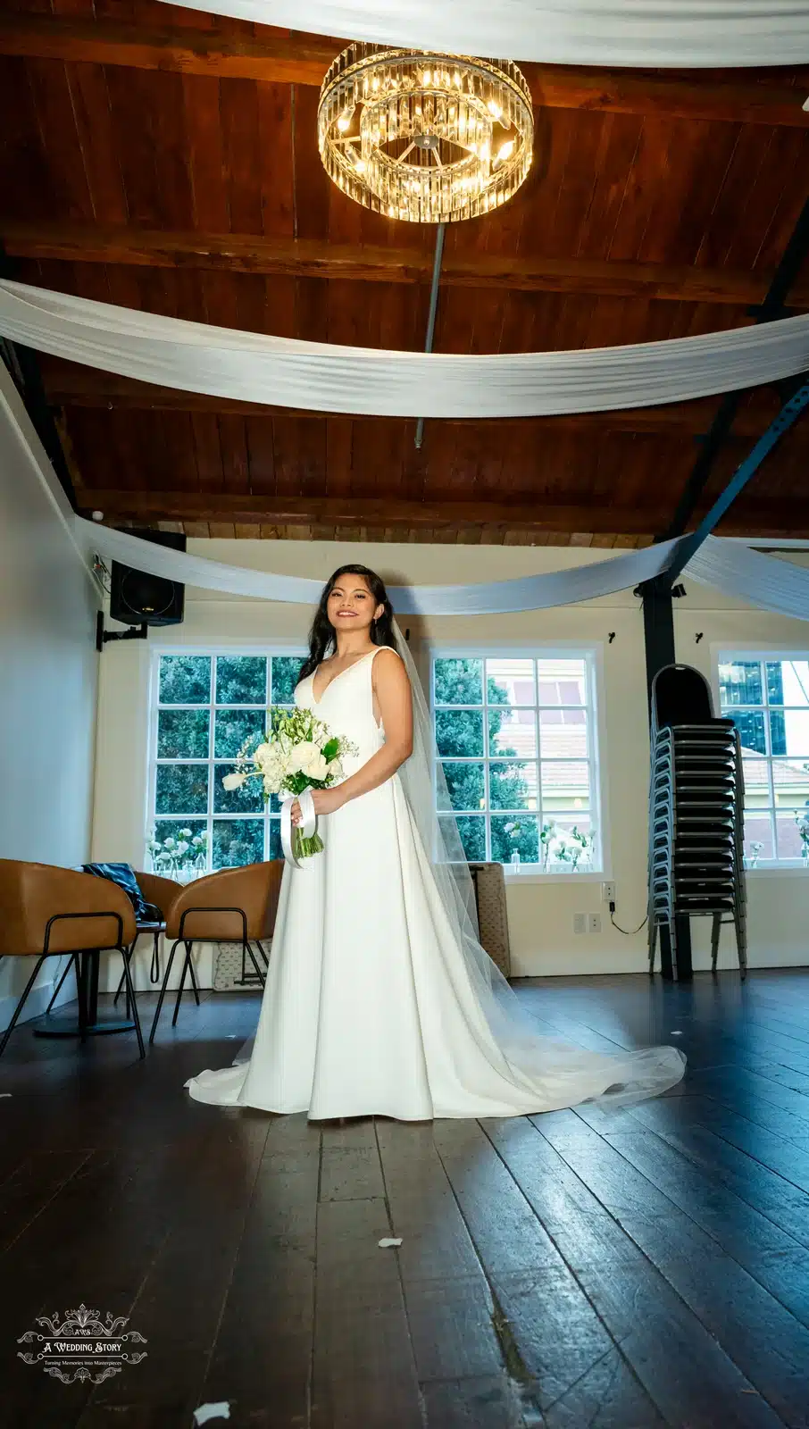 A bride in a classic white gown with a flowing veil stands gracefully under a chandelier in a well-lit indoor venue, holding a bouquet of white flowers, captured beautifully by A Wedding Story.