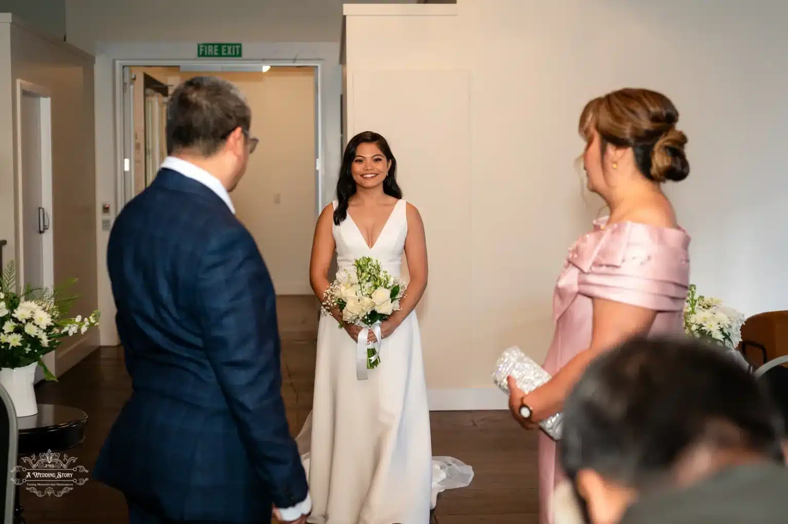 The bride, dressed in a stunning white gown and holding a bouquet of white roses, smiles warmly as she stands facing her parents before the ceremony in Wellington.