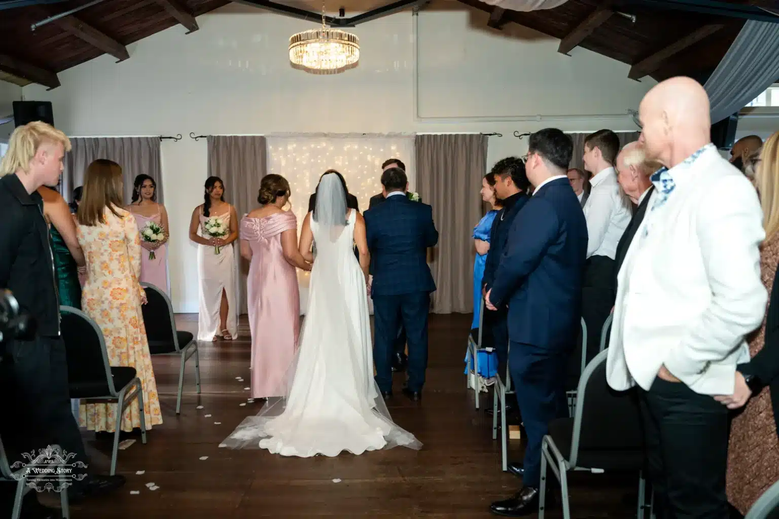 A beautiful bride with a long flowing veil walking down the aisle alongside her parents, surrounded by seated guests, toward a warmly lit altar adorned with fairy lights during a wedding ceremony in Wellington.