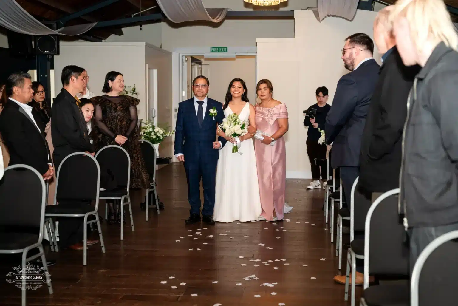 The bride, glowing in her white gown and holding a bouquet of white roses, walks arm-in-arm with her parents down the aisle, surrounded by smiling guests at a Wellington wedding ceremony.