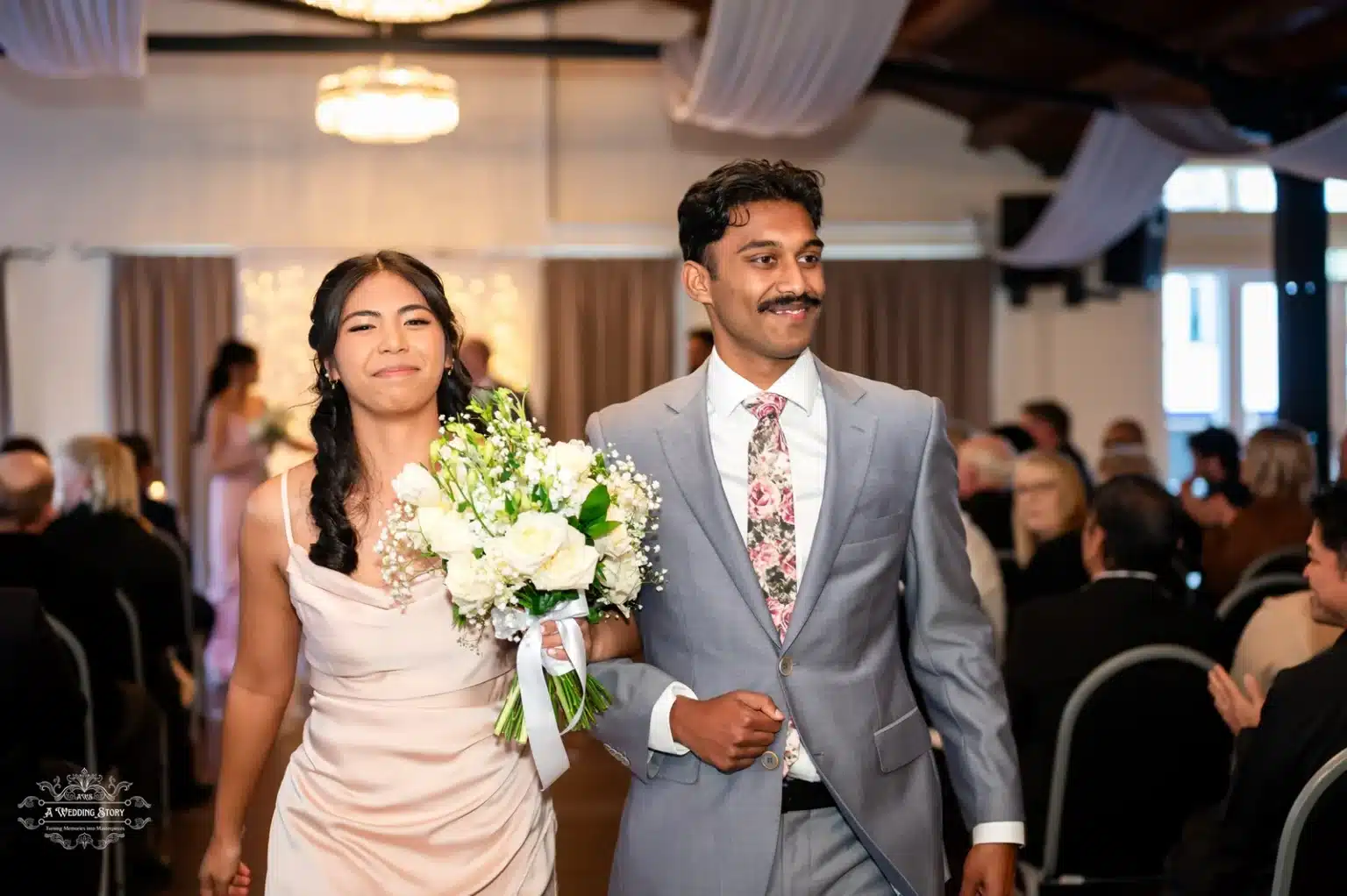 A bridesmaid in a light pink dress and a groomsman in a grey suit with a floral tie walking down the aisle during a wedding ceremony.