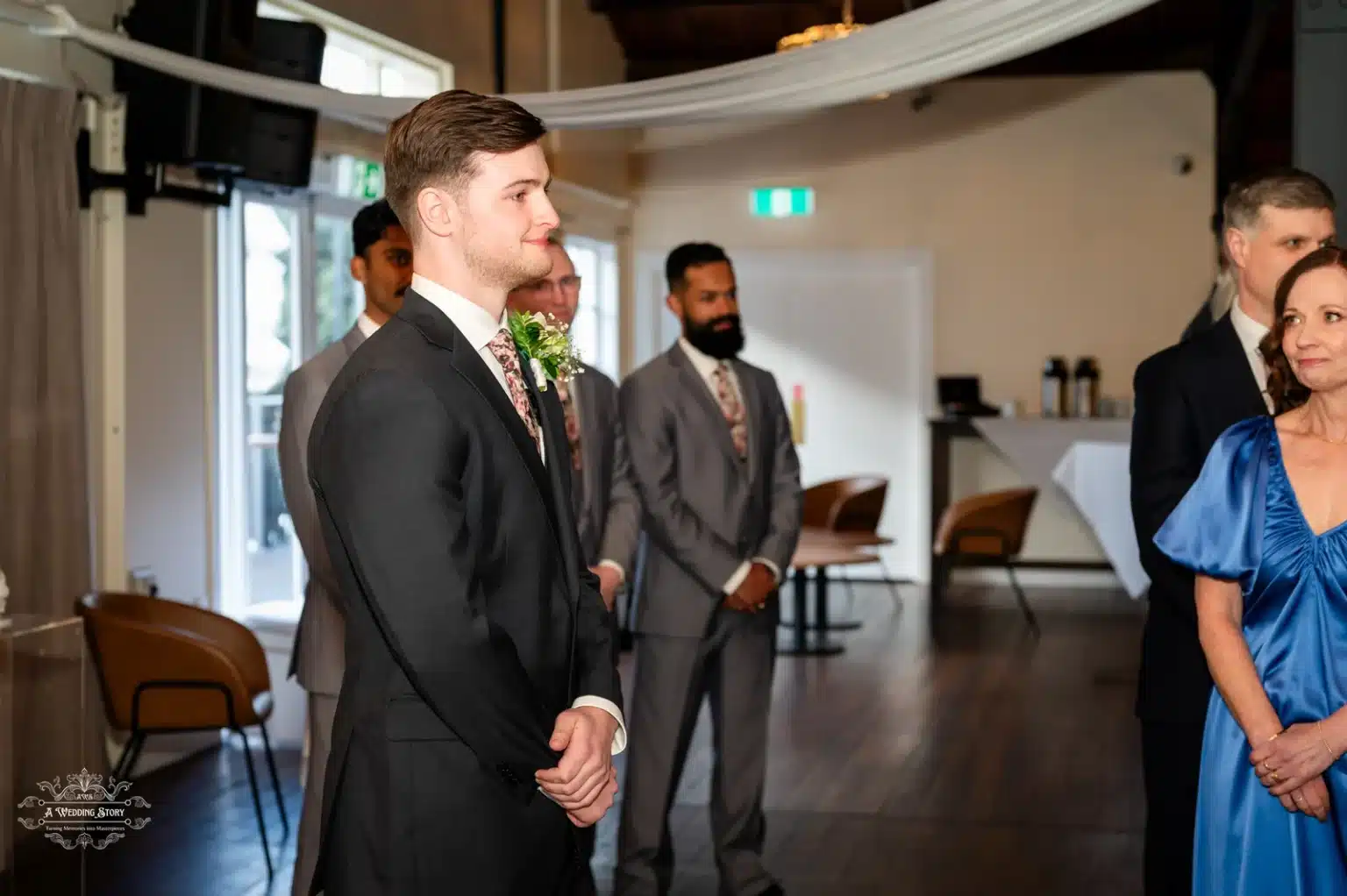 The groom, dressed in an elegant black suit with a floral tie, stands confidently with his groomsmen in the background, eagerly awaiting the bride’s entrance during a wedding ceremony in Wellington.