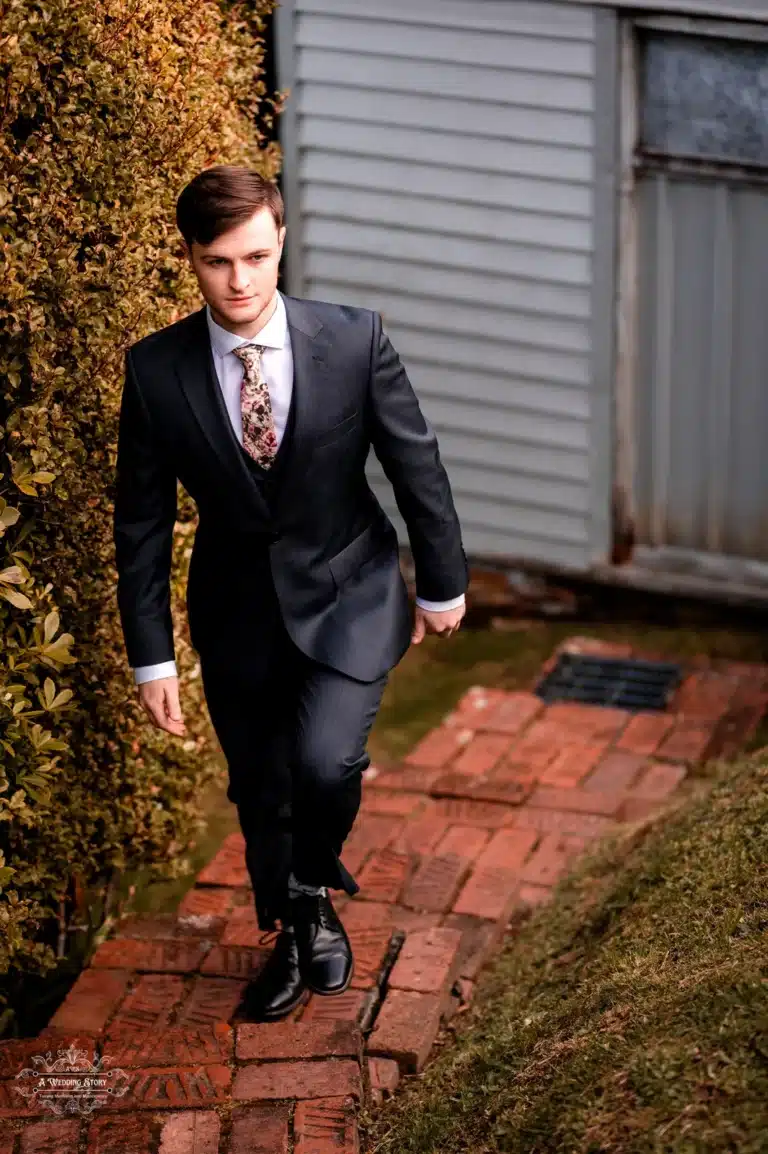 A groom in a tailored navy blue suit with a floral tie walks confidently on a brick pathway, surrounded by greenery, before his wedding ceremony in Wellington, New Zealand.