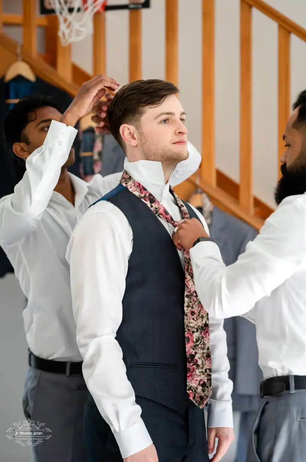 Groom receiving assistance from his groomsmen while getting ready for the wedding day, adjusting his floral tie.