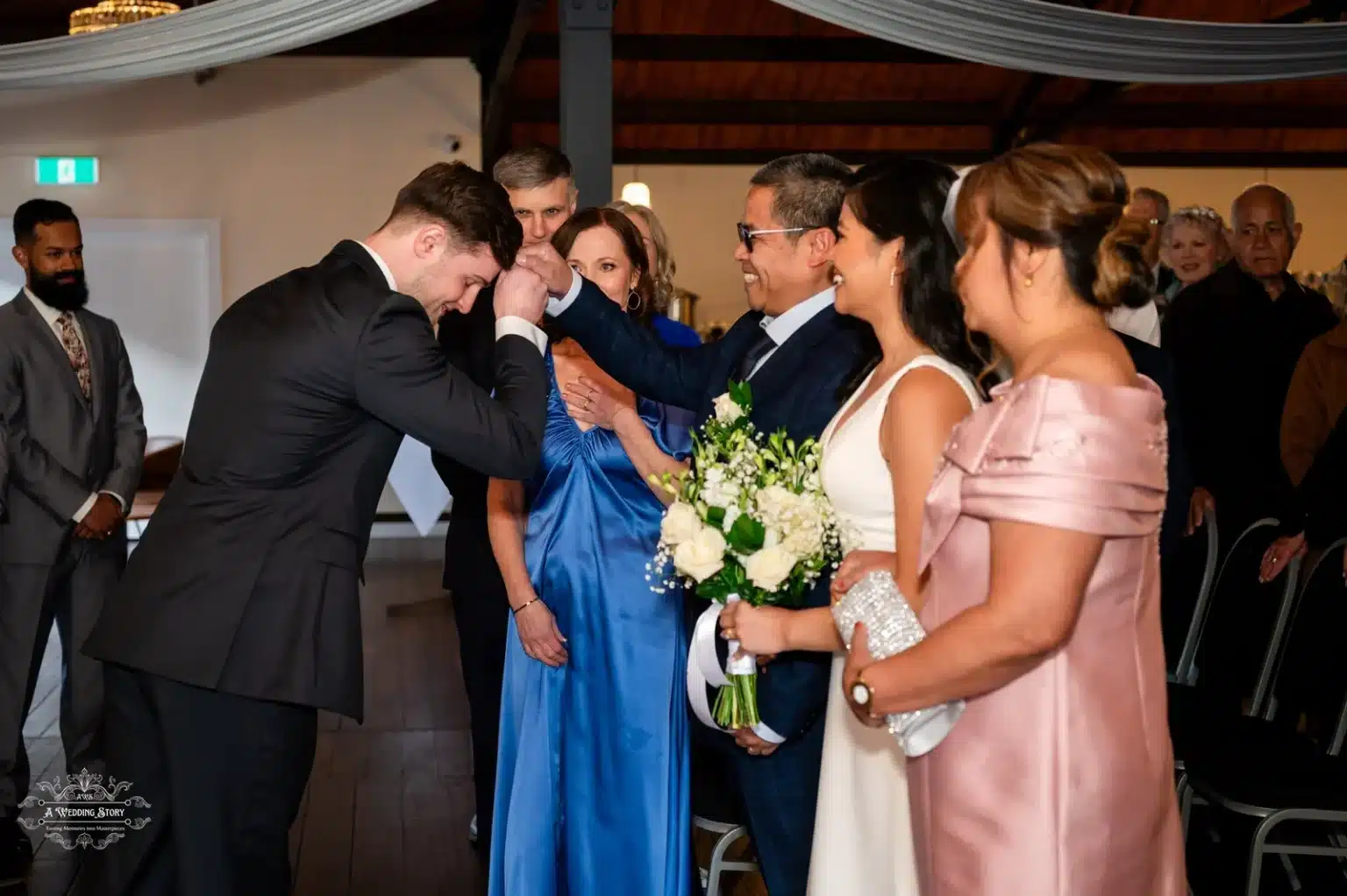 The groom respectfully bows his head as the bride’s father blesses him during an emotional moment at a wedding ceremony in Wellington. The bride, holding a bouquet of white roses, stands beside her parents, beaming with happiness.