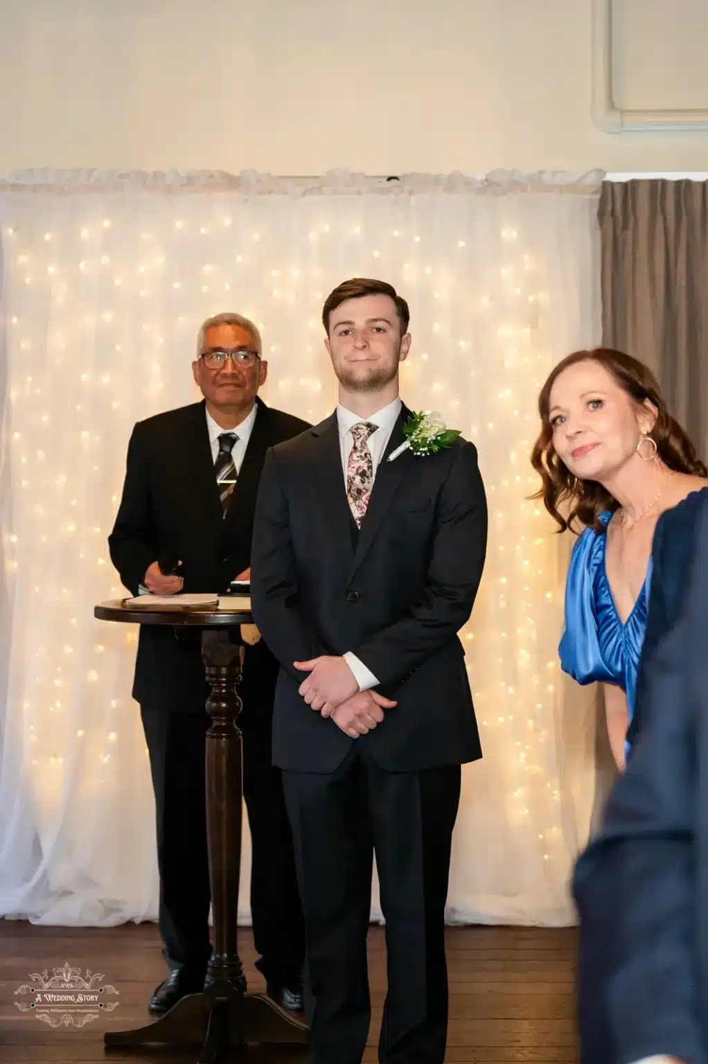 The groom stands confidently with the wedding officiant at a Wellington ceremony, adorned with warm fairy lights as a backdrop. A guest in a vibrant blue dress is visible in the foreground.