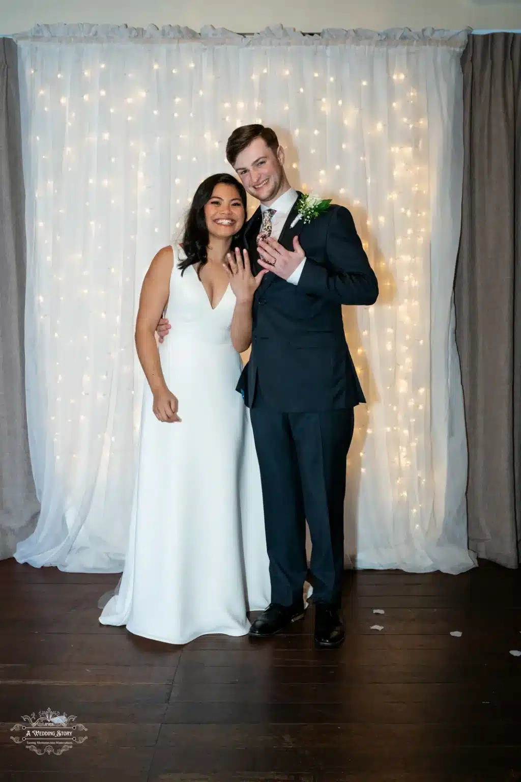 Smiling bride and groom posing with their wedding rings in front of a lighted backdrop in Wellington, New Zealand.