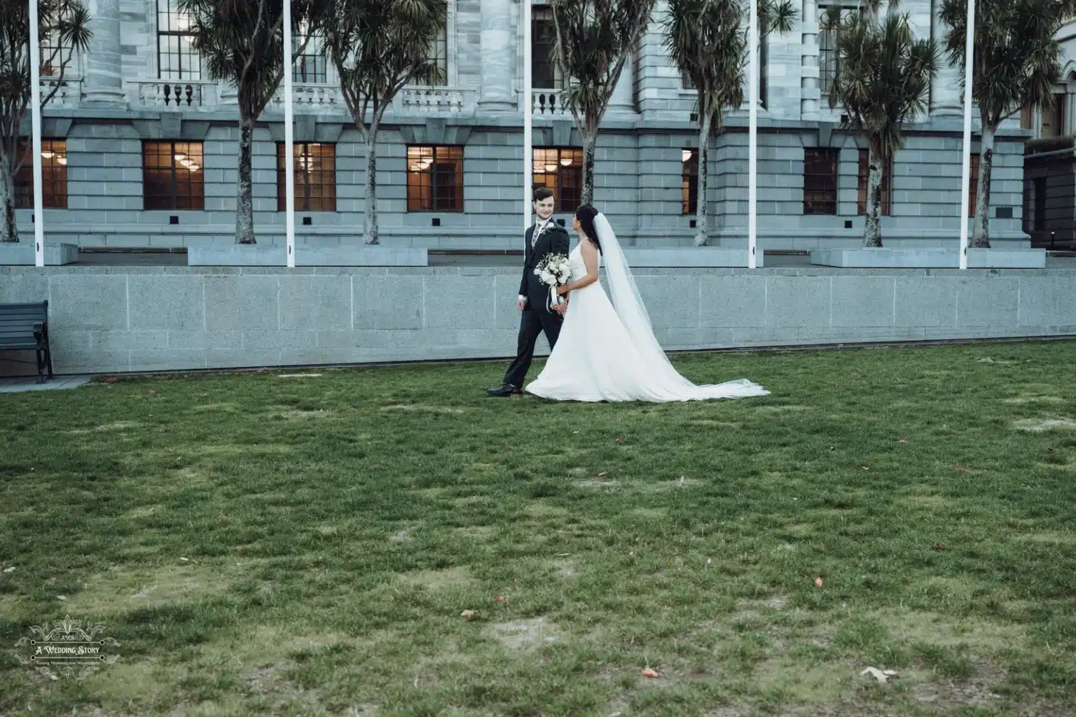 Bride and groom walking gracefully on a lush green lawn with Wellington's historic Parliament buildings in the background.