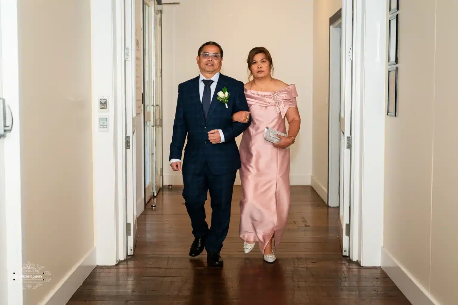 The parents of the bride, dressed elegantly in a navy suit and a blush pink gown, walking arm-in-arm down the aisle during a Wellington wedding ceremony.