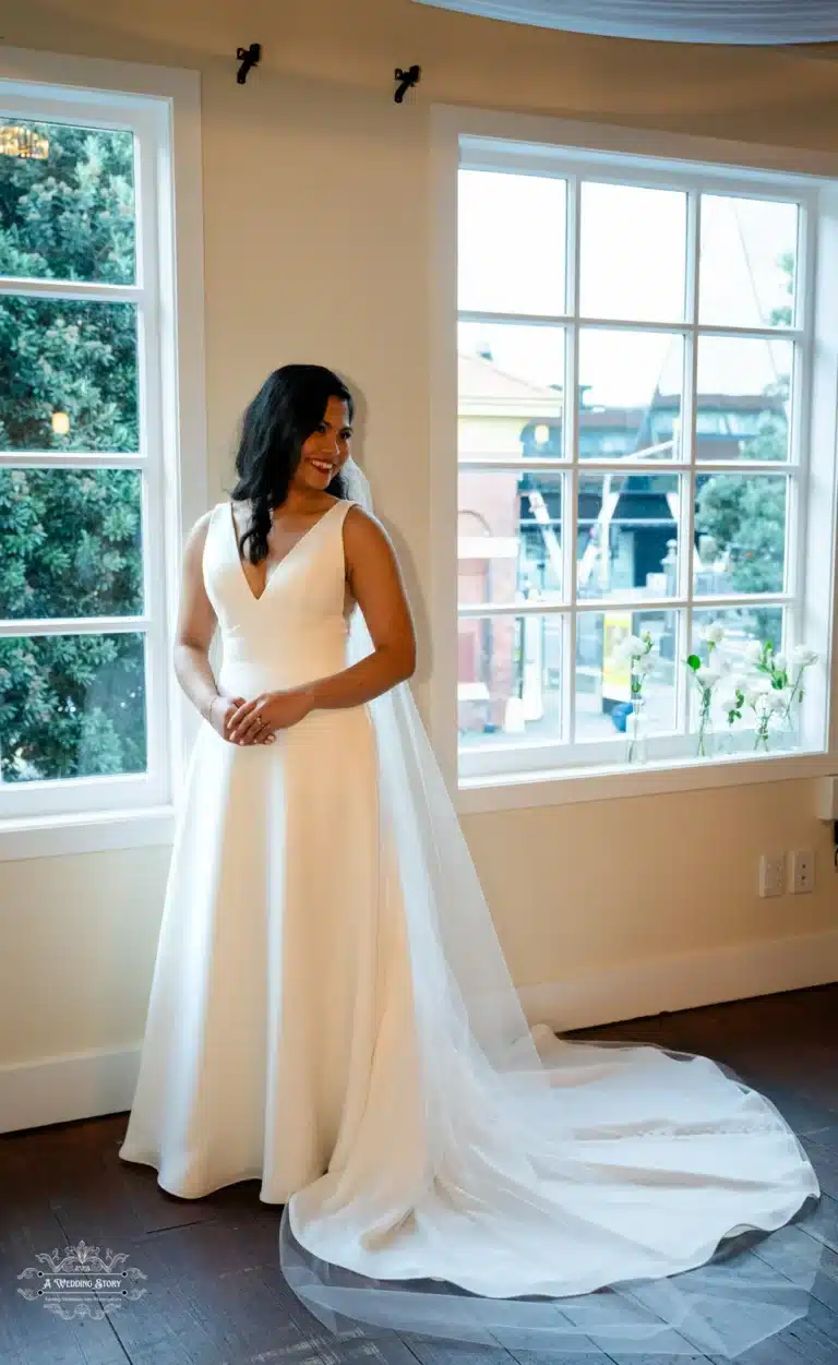 A smiling bride in a white gown with a long veil, standing beside a bright window at a Wellington wedding venue.